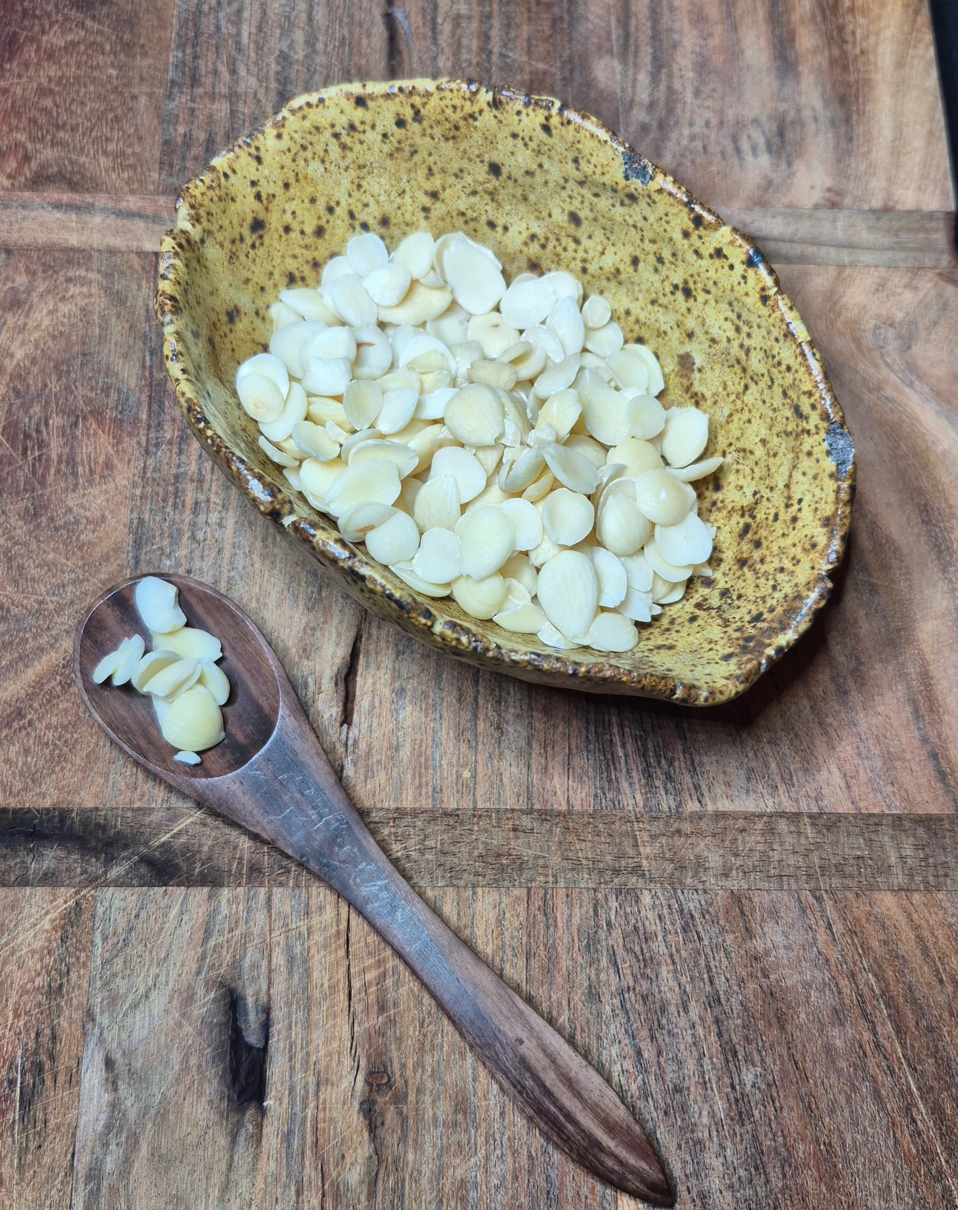 "Close-up of dried apricot kernel on a wooden spoon, with additional herbs in a white ceramic bowl on a rustic wooden surface."